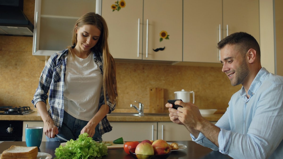 Attractive couple in the kitchen. Man playing video game on smartphone while his girlfriend cooking breakfast early morning at home